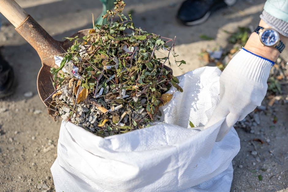 A person wearing a white gardening glove and a wristwatch is using a small shovel to load a white compost bag with garden debris, including cuttings and small branches with green and purple leaves. Th