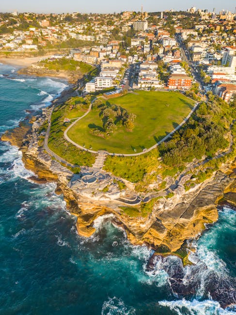 An aerial view of a coastal garden situated on a rocky promontory overlooking the ocean, surrounded by cliffs and with a paved pathway circling the grassy area. The garden features well-maintained lus